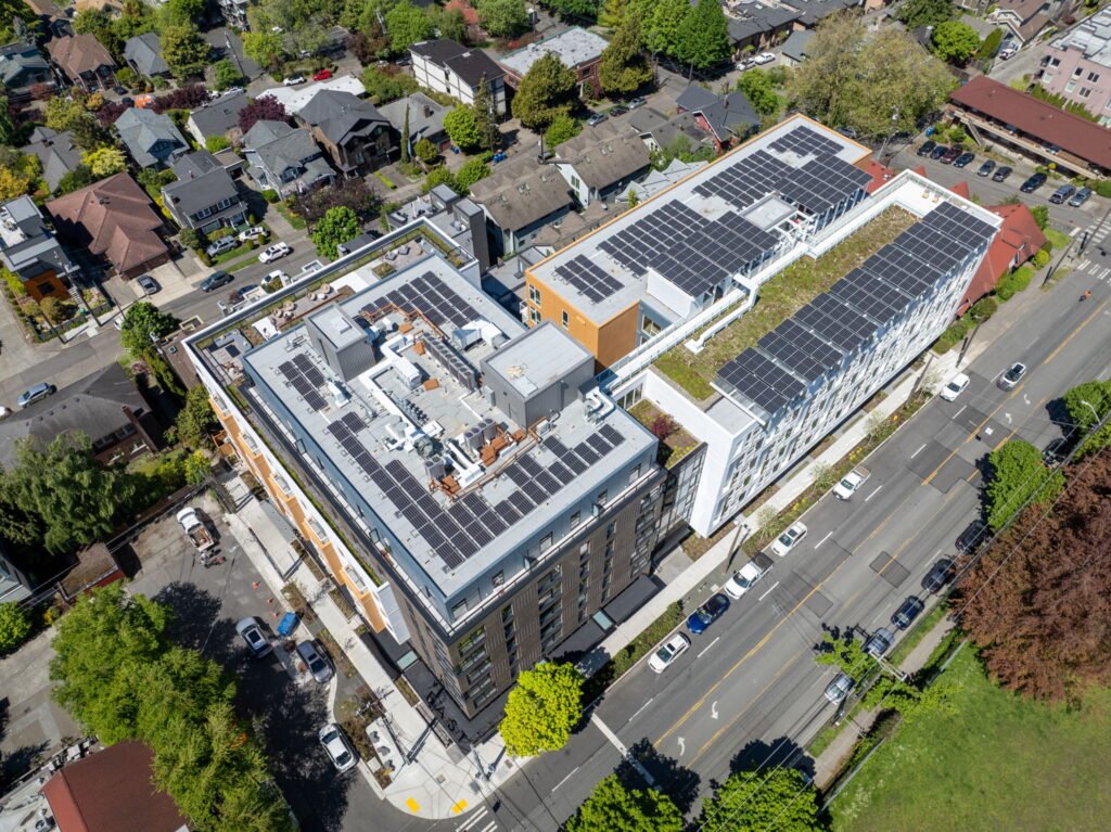 Aerial view of a multi-story building with extensive rooftop solar panels, surrounded by residential streets and green trees in a city block.