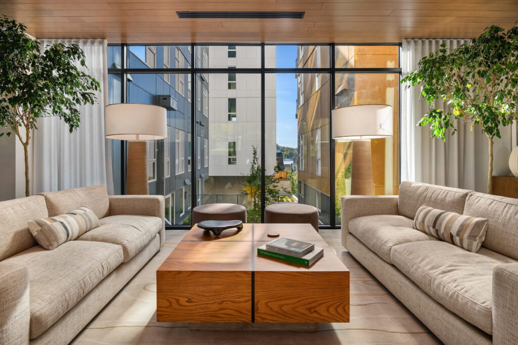 Modern living room with beige sectionals facing a wood coffee table, flanked by tall lamps and large glass walls overlooking neighboring buildings and a blue sky.