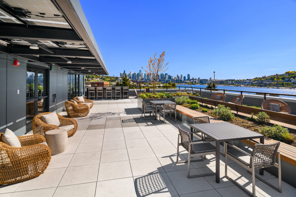 Rooftop terrace with wicker chairs and gray tables, overlooking a city skyline and river under a clear blue sky.