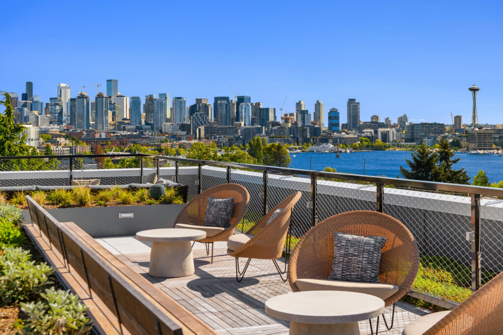 Rooftop terrace with wicker chairs and round concrete tables overlooking a city skyline by the water, Space Needle visible on the right.