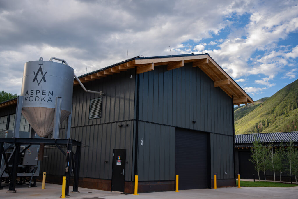 Industrial building with a large silver Aspen Vodka silo beside a dark gray metal warehouse, mountain backdrop.