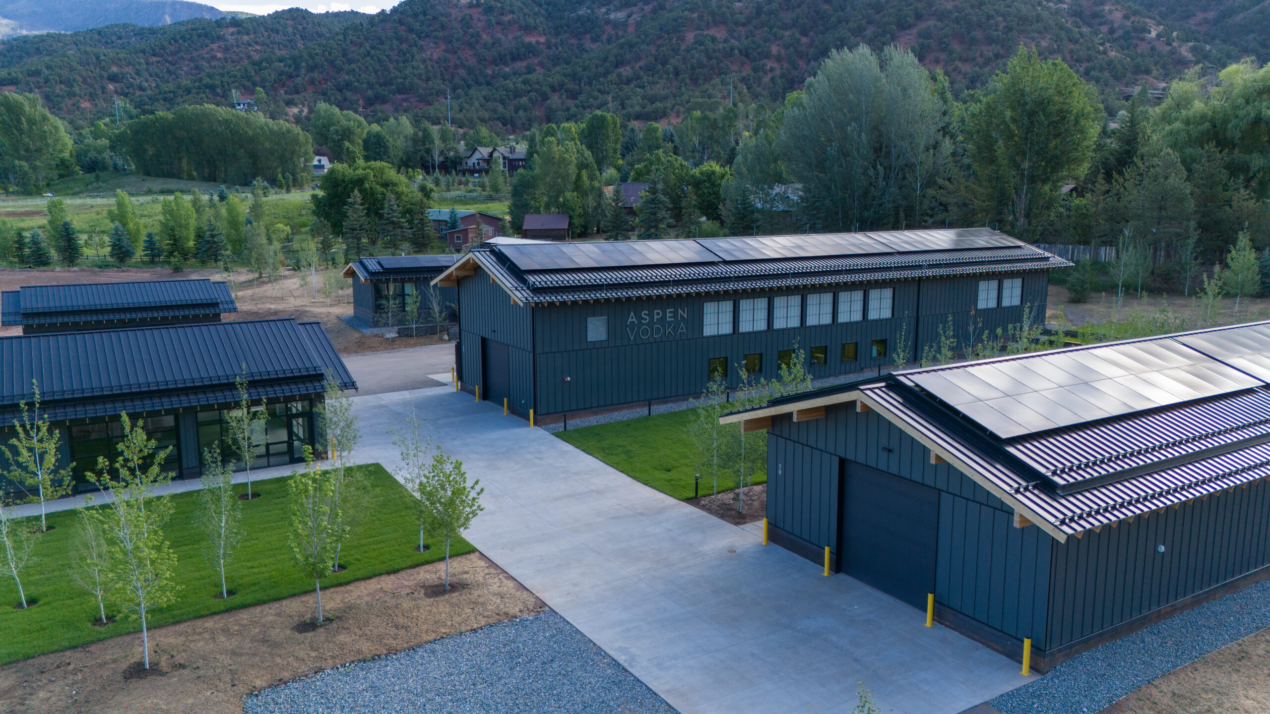 Aerial view of a modern industrial complex with dark gray buildings and solar panels, set against green trees and rolling hills; sign reads 'Aspen Vodka' on the main building
