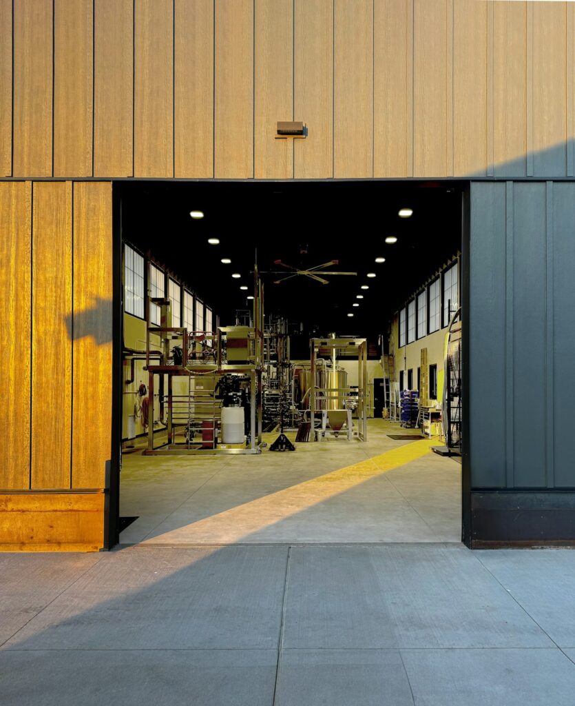 Open industrial workshop doorway showing machinery and scaffolding inside a brightly lit interior, framed by wooden exterior panels bathed in sunlight.