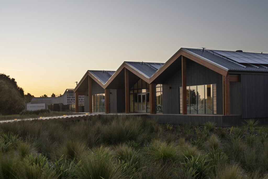 Modern building with a zigzag metal roof and large glass windows at sunset, surrounded by grass and shrubs at ground level.