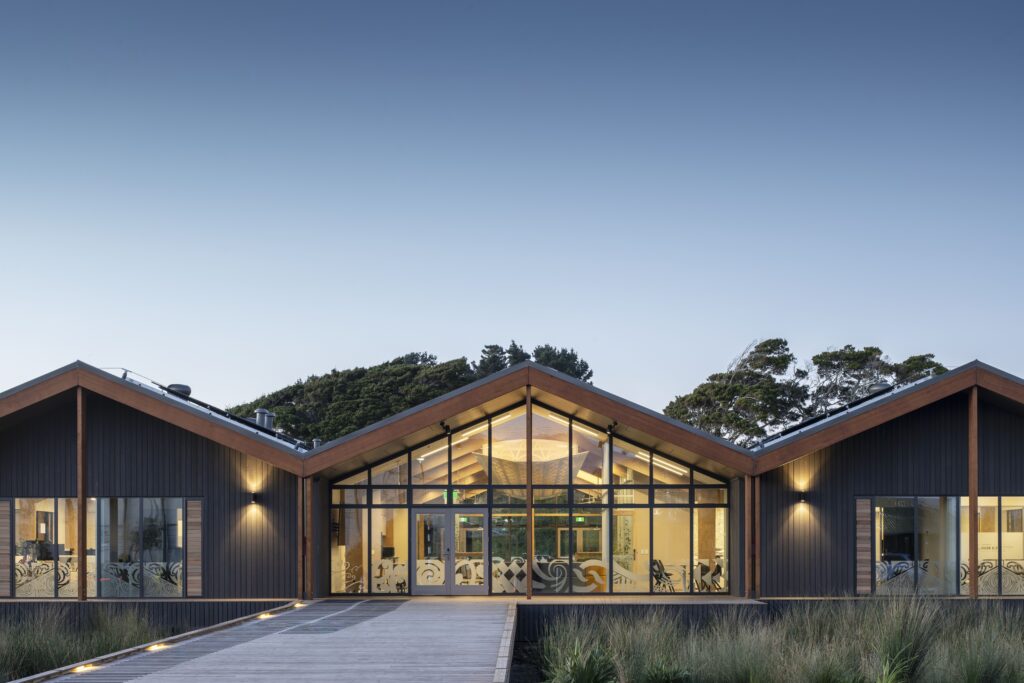 Modern building with wooden beams and large glass entrance, set against a clear sky with surrounding landscaping at dusk