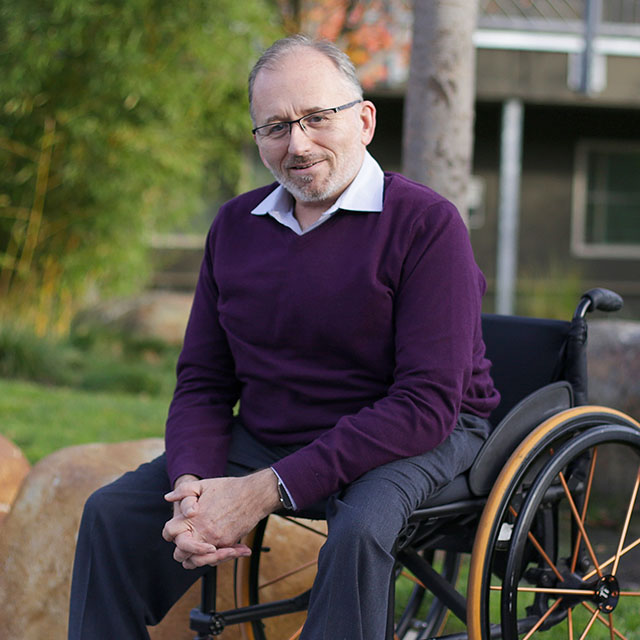 Smiling man in a purple sweater sits in a wheelchair outdoors in a park setting.