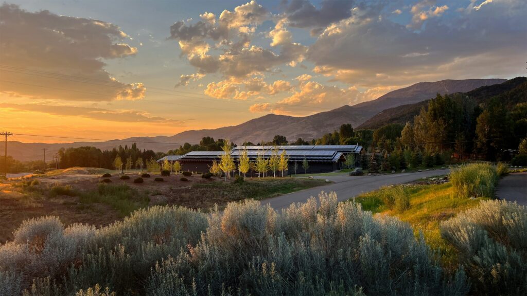 Sunset over a rural landscape with a solar-paneled building, trees, and distant mountains under a warm, cloudy sky.