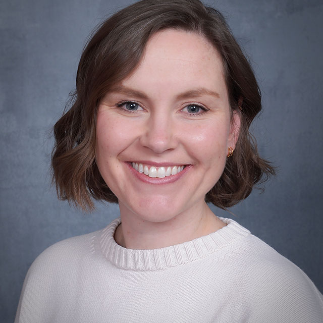 Portrait of a smiling woman with short brown hair and a cream sweater, against a gray backdrop.