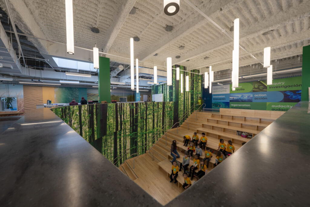 Group of children in yellow shirts seated on wooden bleachers inside a bright science/education space with green walls and banners.