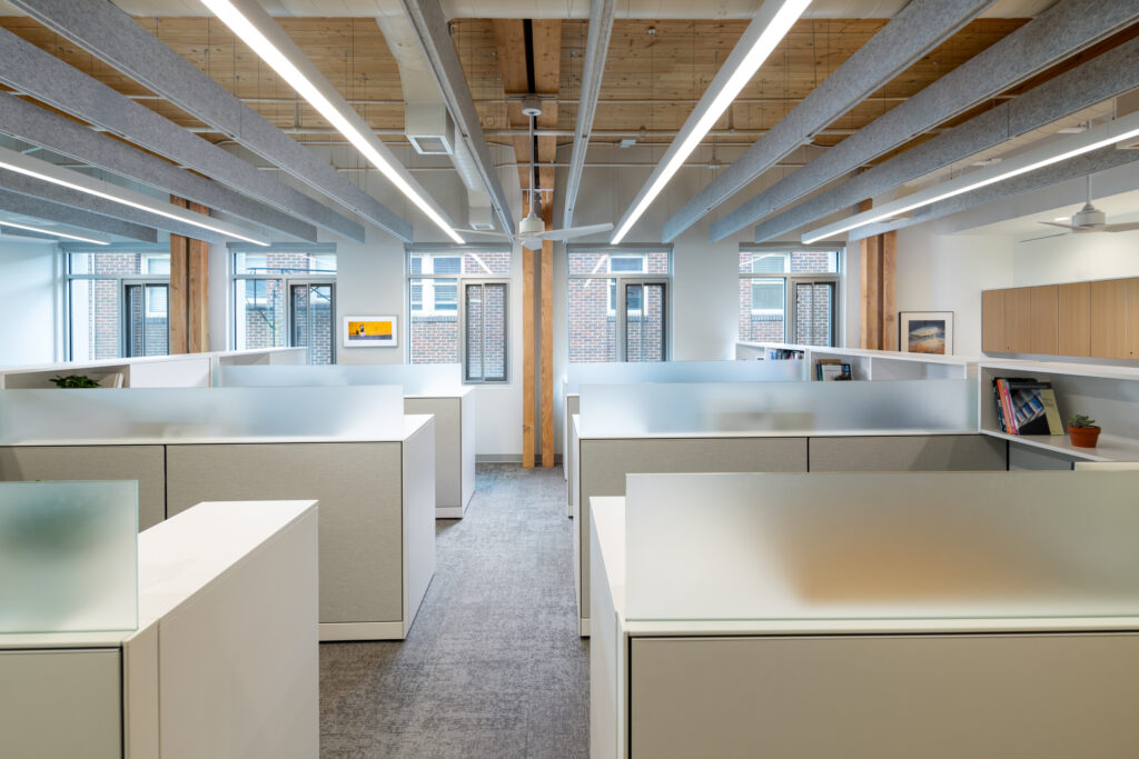 default - Living Future Open-plan office with white cubicles, frosted glass dividers, and exposed wood ceiling beams.