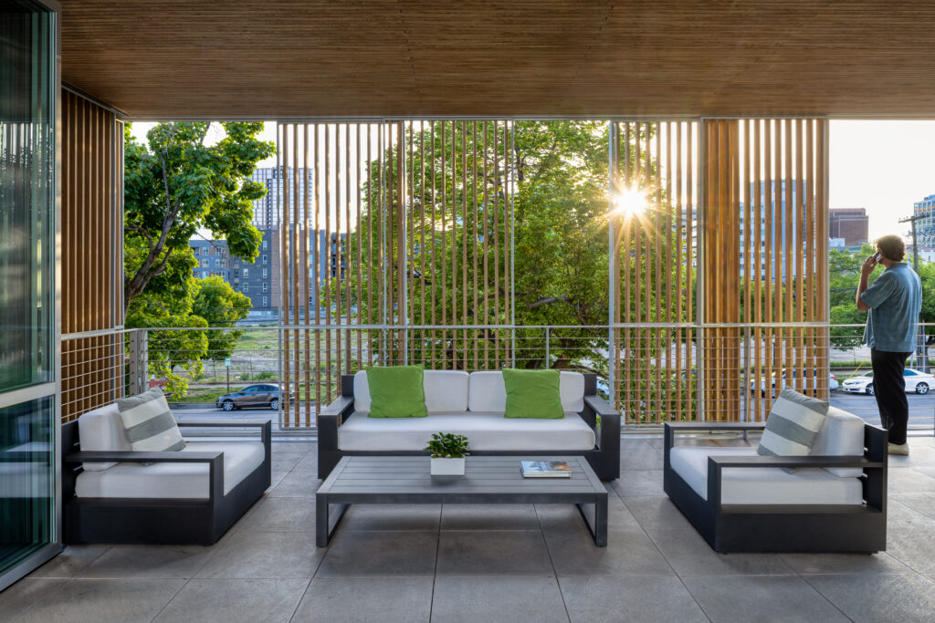 default - Living Future Outdoor balcony lounge with white-cushioned sectional sofas and green pillows, wooden slat screens, and a sunset behind trees in the cityscape.