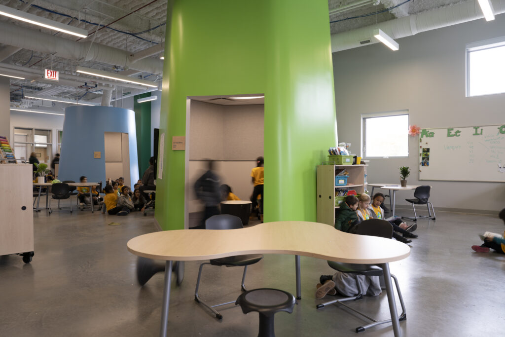 Children sit on the floor and desks around rounded tables in a bright classroom with a tall green pillar and a whiteboard on the right.