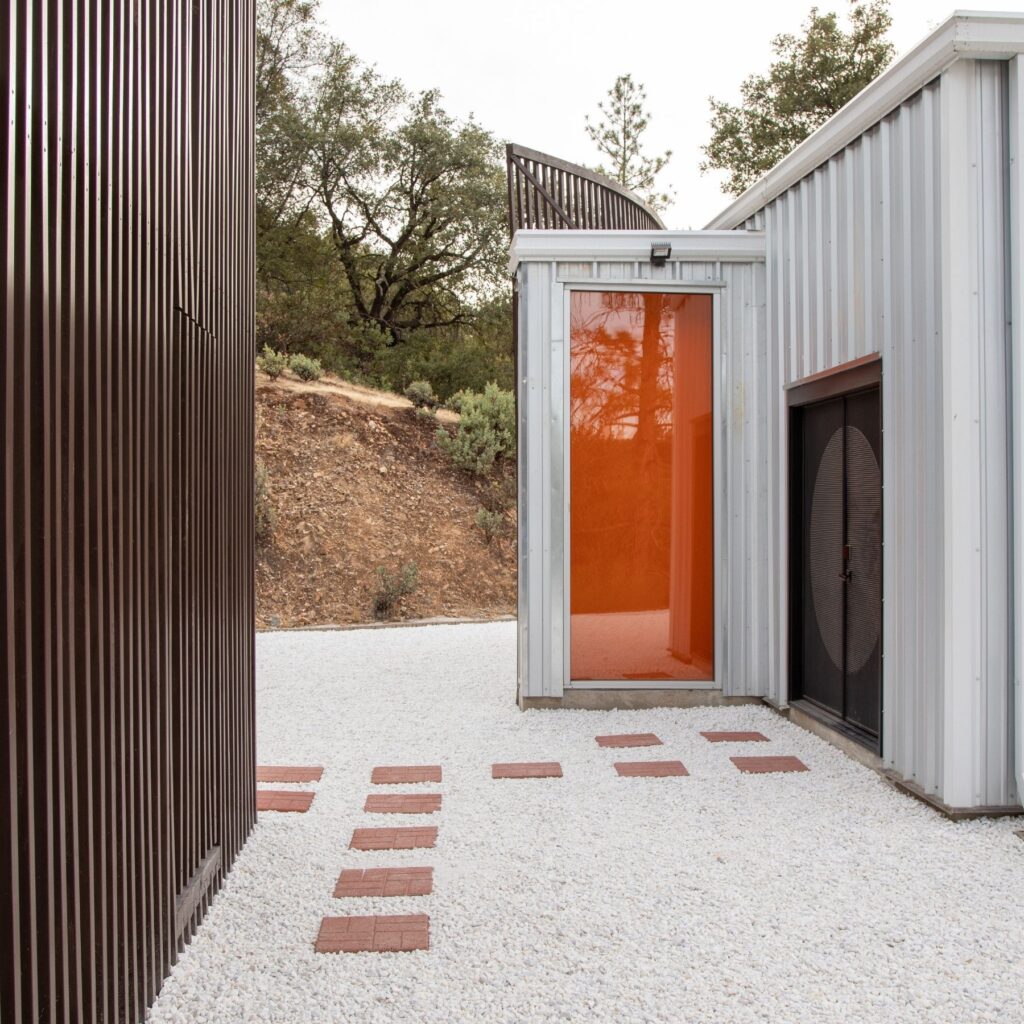 BoxFactory_2 - Living Future Narrow courtyard between dark brown slatted wall and metal-clad building, with a stepping-stone path on white gravel.