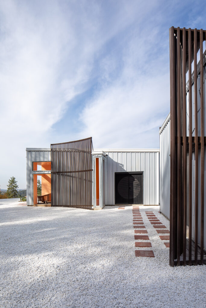 QuiteOak--14 - Living Future Modern building facade with vertical brown slats, orange-framed entryways, and a gravel yard under a blue sky.