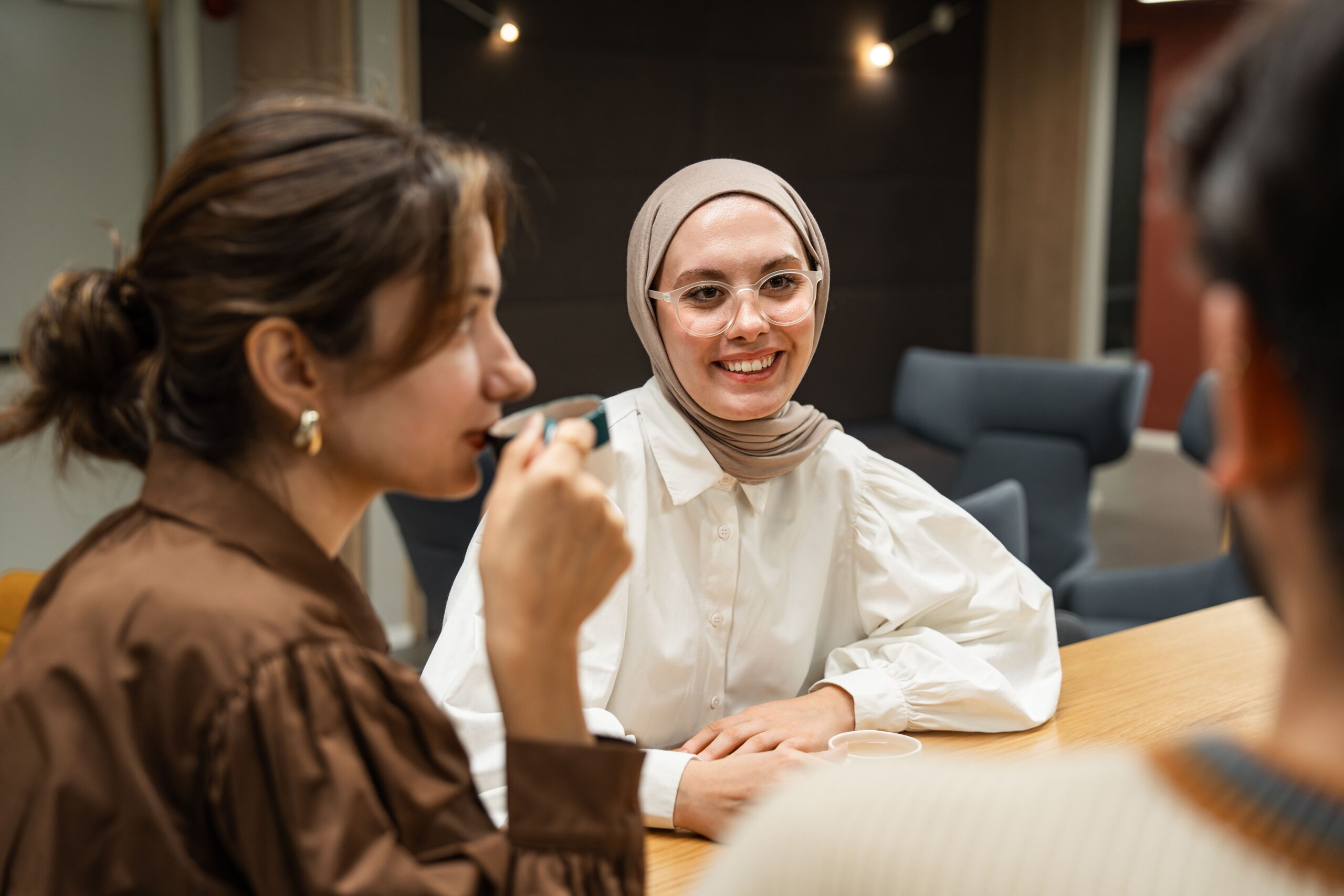 pexels-mutecevvil-20450974 - Living Future Woman in a beige hijab and glasses smiles while chatting with colleagues at a wooden table in a modern meeting room.
