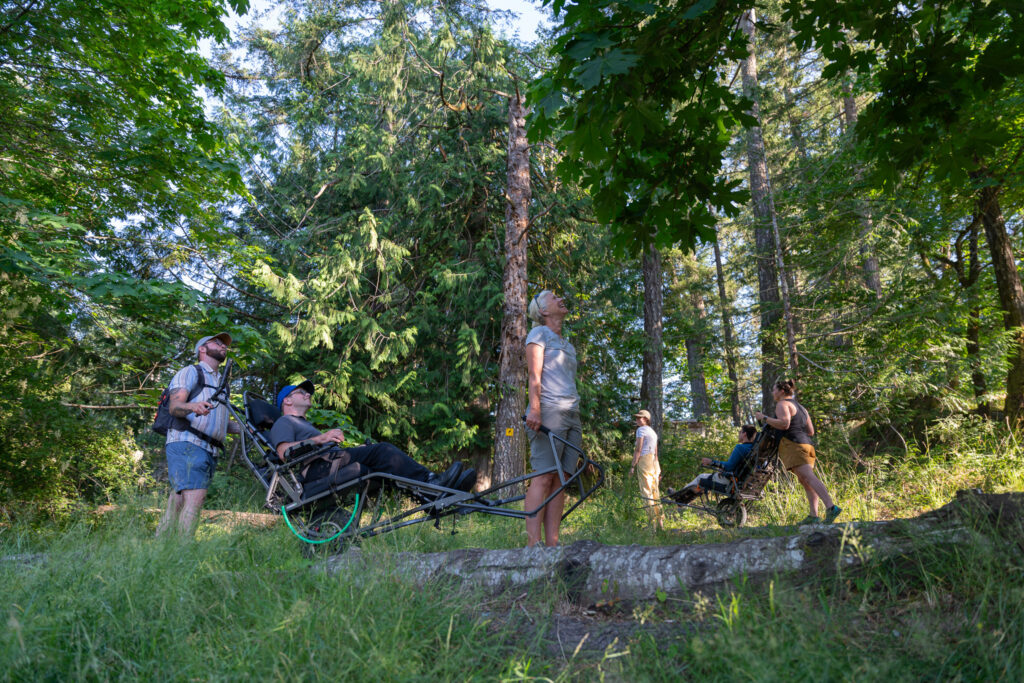 Group of people hiking in a green forest; one person uses an adaptive wheelchair bike while others have bicycles nearby.