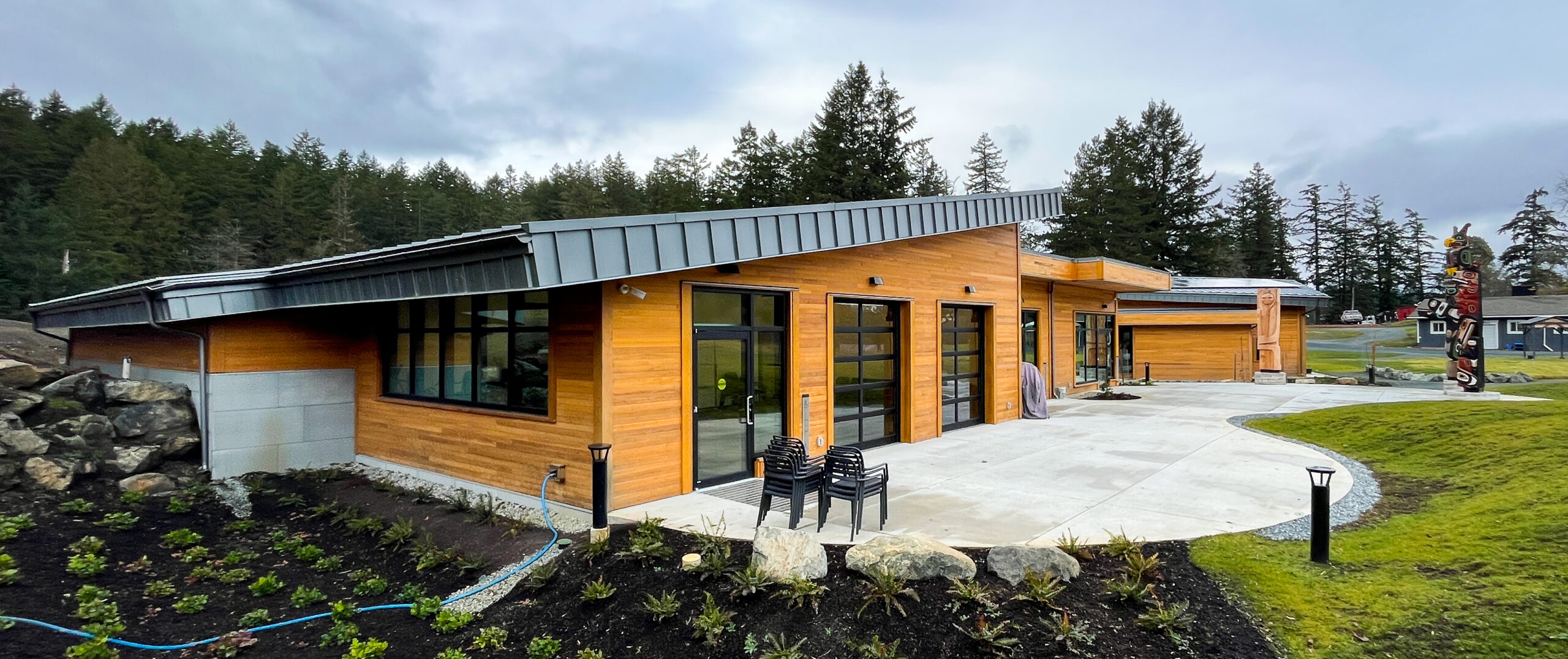 Modern wooden building with large glass doors and a row of stacked chairs outside, set against a wooded backdrop and a curved concrete path.