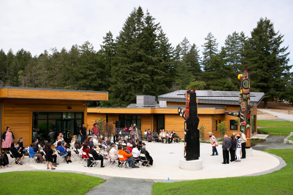 Outdoor ceremony or performance with rows of seated attendees in a courtyard, wooden building behind, colorful totem poles on the right, and pine trees in the background.