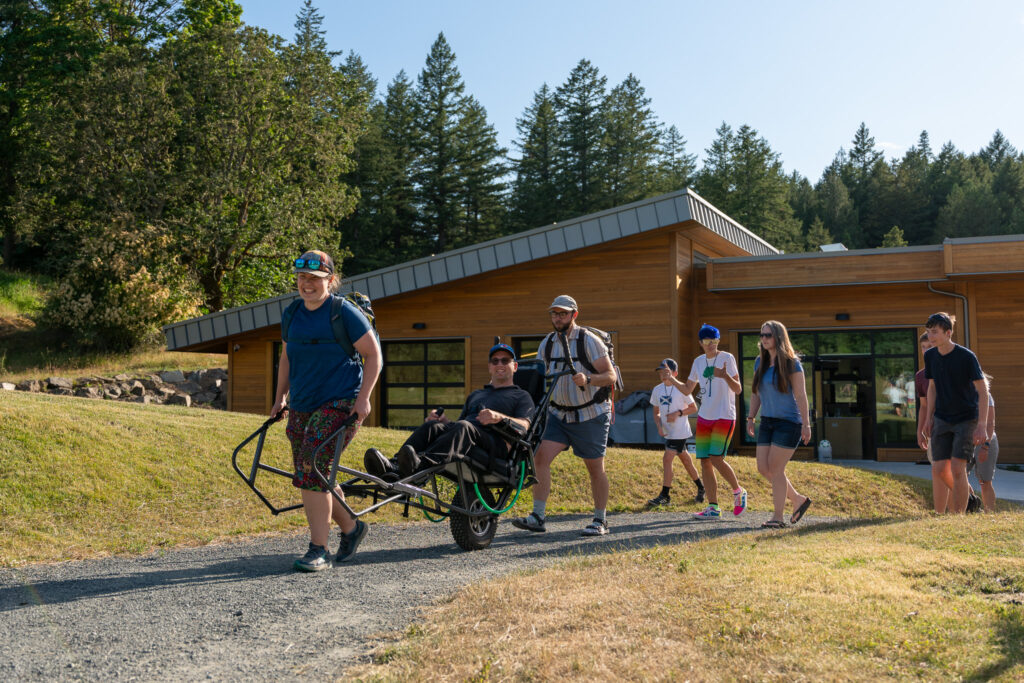 Group of people walking outdoors in front of a wood-clad building with trees in the background; one person uses a powered wheelchair accompanied by others on a sunny day.