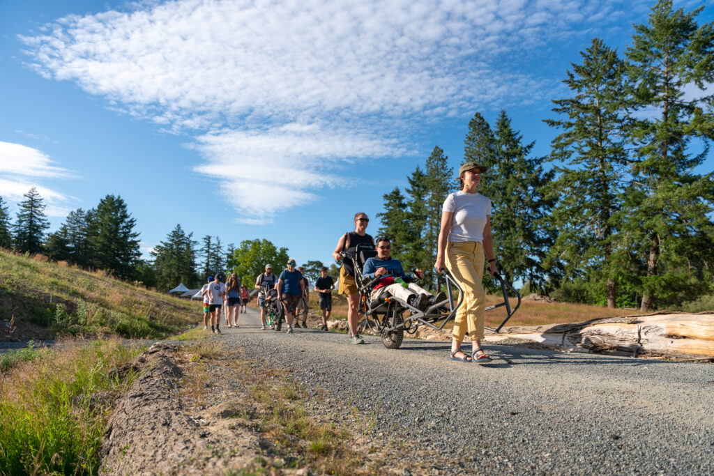 Group of people walking on a gravel trail in a sunny park, with a woman in a white shirt pushing a person in a wheelchair at the front and pine trees in the background.