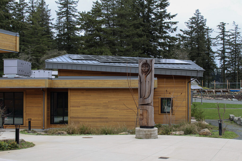 Wood-clad building with a metal roof and solar panels, plus a carved wooden sculpture in the foreground.