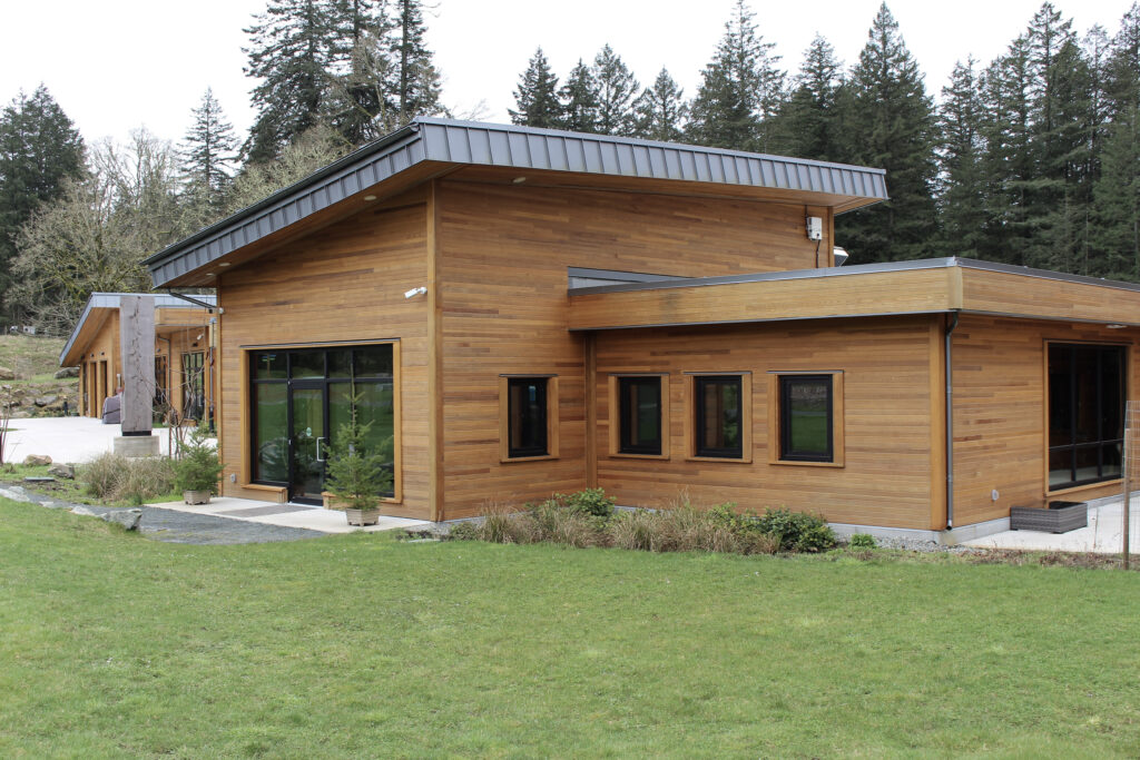 Wood-clad modern building with large windows and sliding glass doors, set on a green lawn with a forest backdrop.