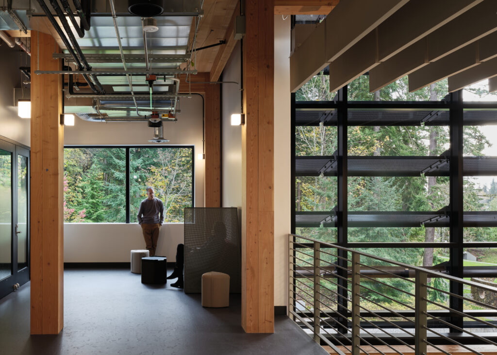 cKevinScott-KaiserBorsariHall0698 - Living Future A man standing in a modern open-concept office with wooden columns, large windows, and a metal railing on the right.