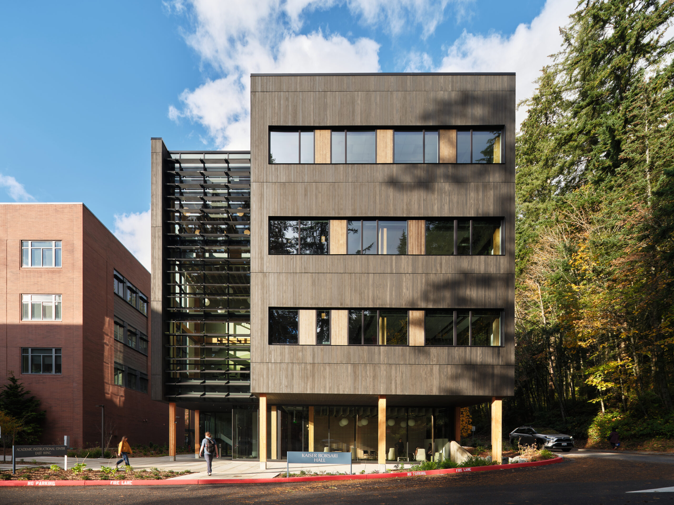 cKevinScott-KaiserBorsariHall1040 - Living Future Modern multi-story building with a wood-clad facade, horizontal windows, and a glass atrium on the left against a blue sky.
