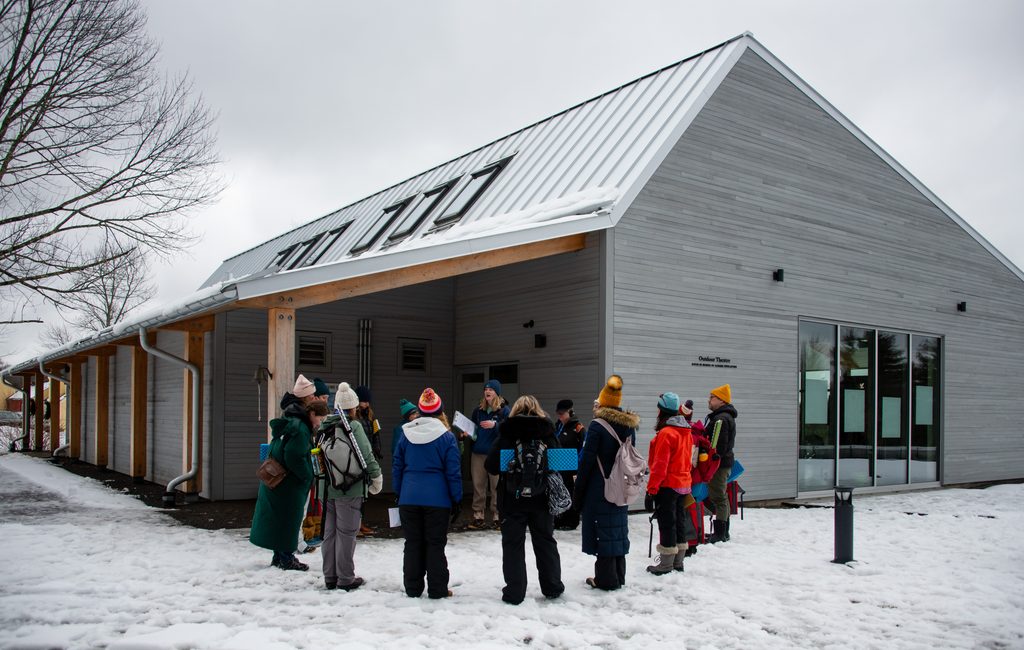Group of winter-clad people gathered outside a gray wooden building with a slanted metal roof and skylights, snow on the ground outside a theatre entrance.