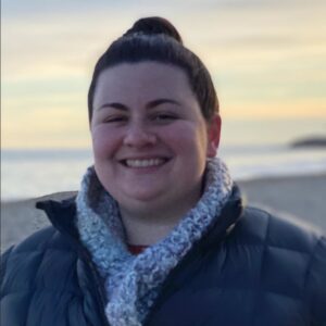 Smiling woman in a bun, wearing a navy puffer jacket and scarf, on a beach at sunset.